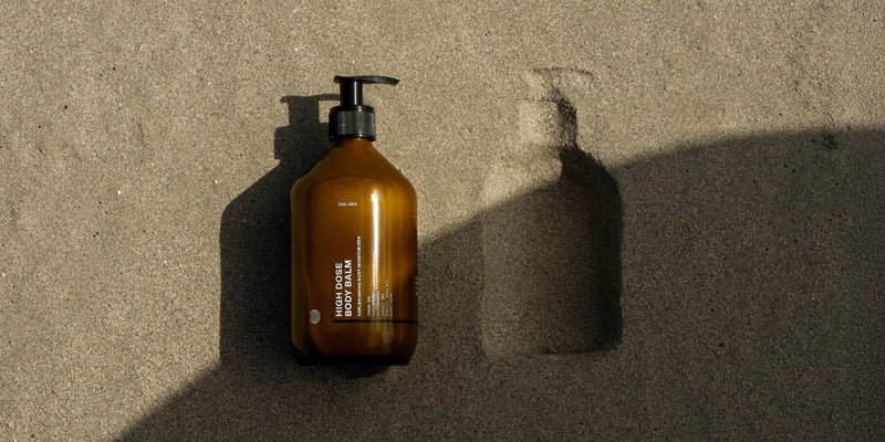 Eine braune Pumpflasche liegt auf sandigem Boden, wirft einen Schatten und hinterlässt einen Abdruck ihrer Form im Sand neben ihr. Das Sonnenlicht hebt die Textur des Sandes und der Flasche hervor.