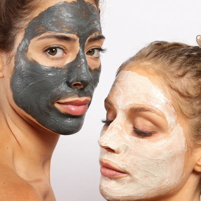Zwei Frauen verwenden das DALUMA 2x MASK BUNDLE: eine trägt eine schwarze Kohlemaske und blickt in die Kamera, während die andere mit einer weißen Tonmaske nach unten blickt, beide vor einem hellen, schlichten Hintergrund.
