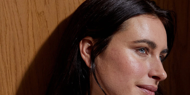 Eine Frau mit langen dunklen Haaren und heller Haut ist in Großaufnahme zu sehen, die nachdenklich zur Seite blickt. Der Hintergrund ist eine hölzerne Fläche, und weiches Licht beleuchtet ihr Gesicht.