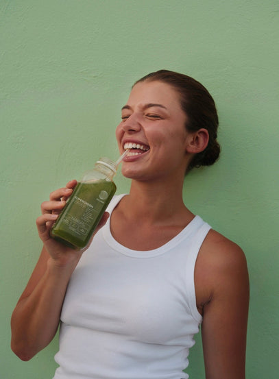 Eine lächelnde Frau in einem weißen Tank-Top hält eine Flasche grünen Saft in der Hand und lacht, während sie vor einer hellgrünen Wand steht.