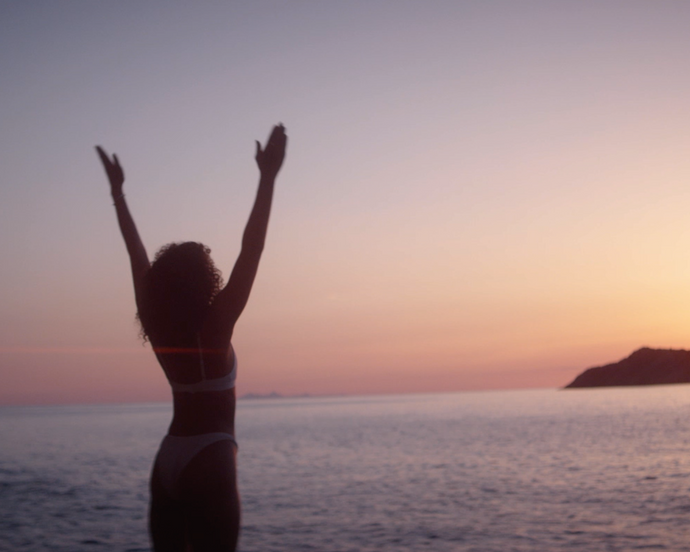 Frau im Bikini steht mit erhobenen Armen und blickt auf ein ruhiges Meer bei Sonnenuntergang. Im Hintergrund sind schemenhafte Berge und ein bunter Himmel zu sehen, die eine friedliche, fröhliche Szene schaffen.