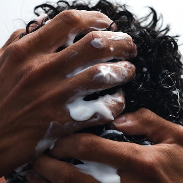 Nahaufnahme von Händen, die weißes Shampoo oder Conditioner in lockiges, nasses Haar einmassieren, wobei die Finger gespreizt sind und der Schaum das Haar und die Hände bedeckt.