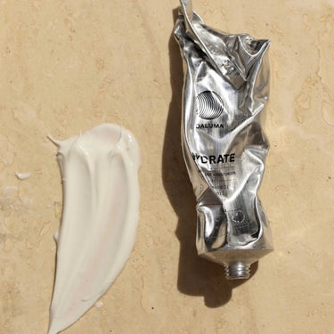Eine zerknitterte silberne Tube mit der Aufschrift HYCRATE von DALUMA's FACIAL MOISTURIZER GESICHTSPFLEGE ruht auf einer beigen Fläche neben einem weißen Cremeschmier und deutet auf eine vegane Feuchtigkeitscreme für empfindliche Haut hin.