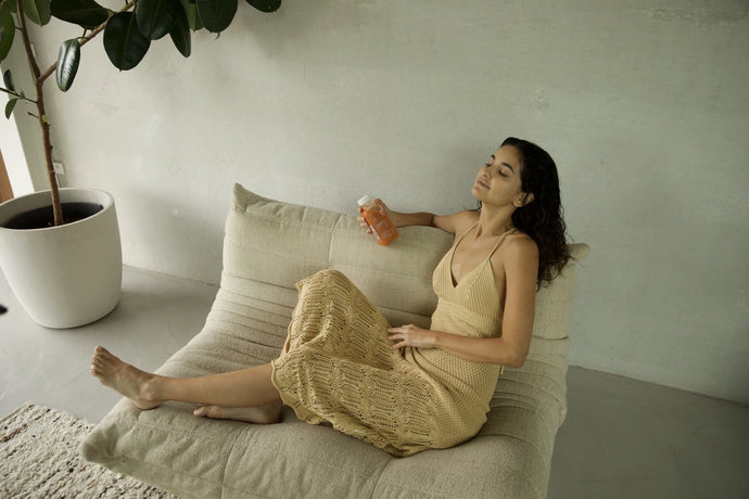Eine Frau in einem hellbeigen Kleid entspannt sich auf einem gepolsterten Sitz und hält mit geschlossenen Augen ein Getränk in ihrer linken Hand. Im Hintergrund ist eine Topfpflanze vor einer hellen Wand zu sehen.