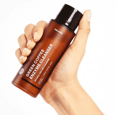 Eine Hand hält eine braune Flasche mit der Aufschrift DALUMA GREEN COFFEE CLEANSER ENZYMPEELING mit schwarzem Verschluss und minimalistischem Design, ideal für sanftes Peeling und natürliche Regeneration, vor einem weißen Hintergrund.