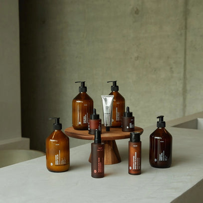 Eine Sammlung von bernsteinfarbenen und braunen Pflegeflaschen und eine silberne Tube sind auf und um einen Holzsockel vor einem minimalistischen Betonhintergrund angeordnet.