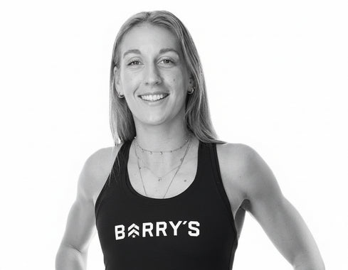 Eine lächelnde Frau mit langen blonden Haaren trägt ein schwarzes Tank-Top, auf dem in weißen Buchstaben BARRY'S steht. Sie steht vor einem schlichten weißen Hintergrund.