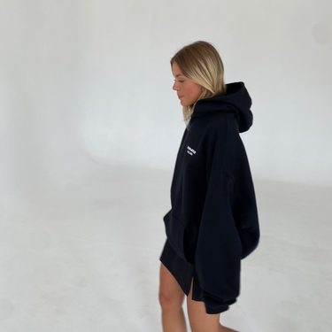 Eine blonde Frau trägt den JOSEPH ATELIER X DALUMA REHAB HOODIE von DALUMA in Oversize-Passform mit weißen Turnschuhen, während sie in einem Haus vor einem schlichten weißen Hintergrund spazieren geht.