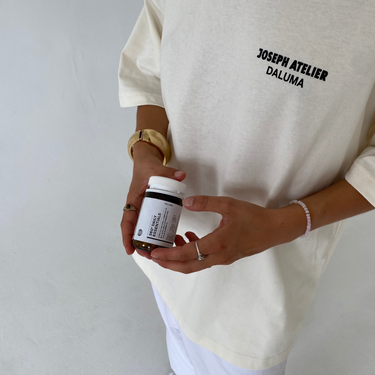 Eine Person in einem weißen JOSEPH ATELIER X DALUMA SELFCARE SUNDAY TEE und einer weißen Hose hält ein kleines Glas mit einem weißen Etikett vor einem schlichten weißen Hintergrund. Marke: DALUMA.