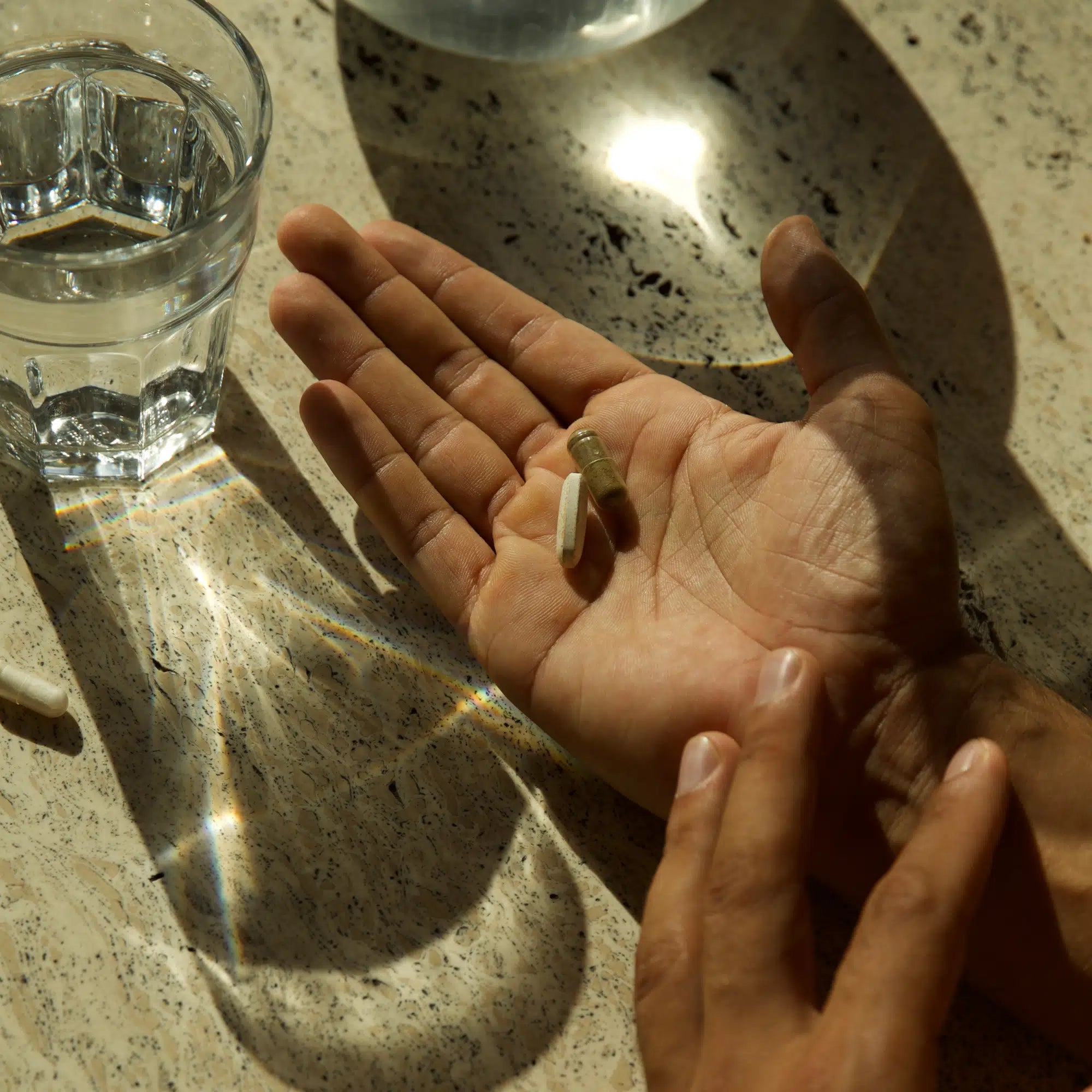 Eine Person hält drei IMMUN DEFENCE COMPLETE KOMPLEX DOUBLE Kapseln von DALUMA in einer Hand, während ein Glas Wasser in der Nähe auf einer sonnenbeschienenen, gesprenkelten Arbeitsplatte das Licht reflektiert und ein Regenbogenmuster auf der Oberfläche erzeugt.