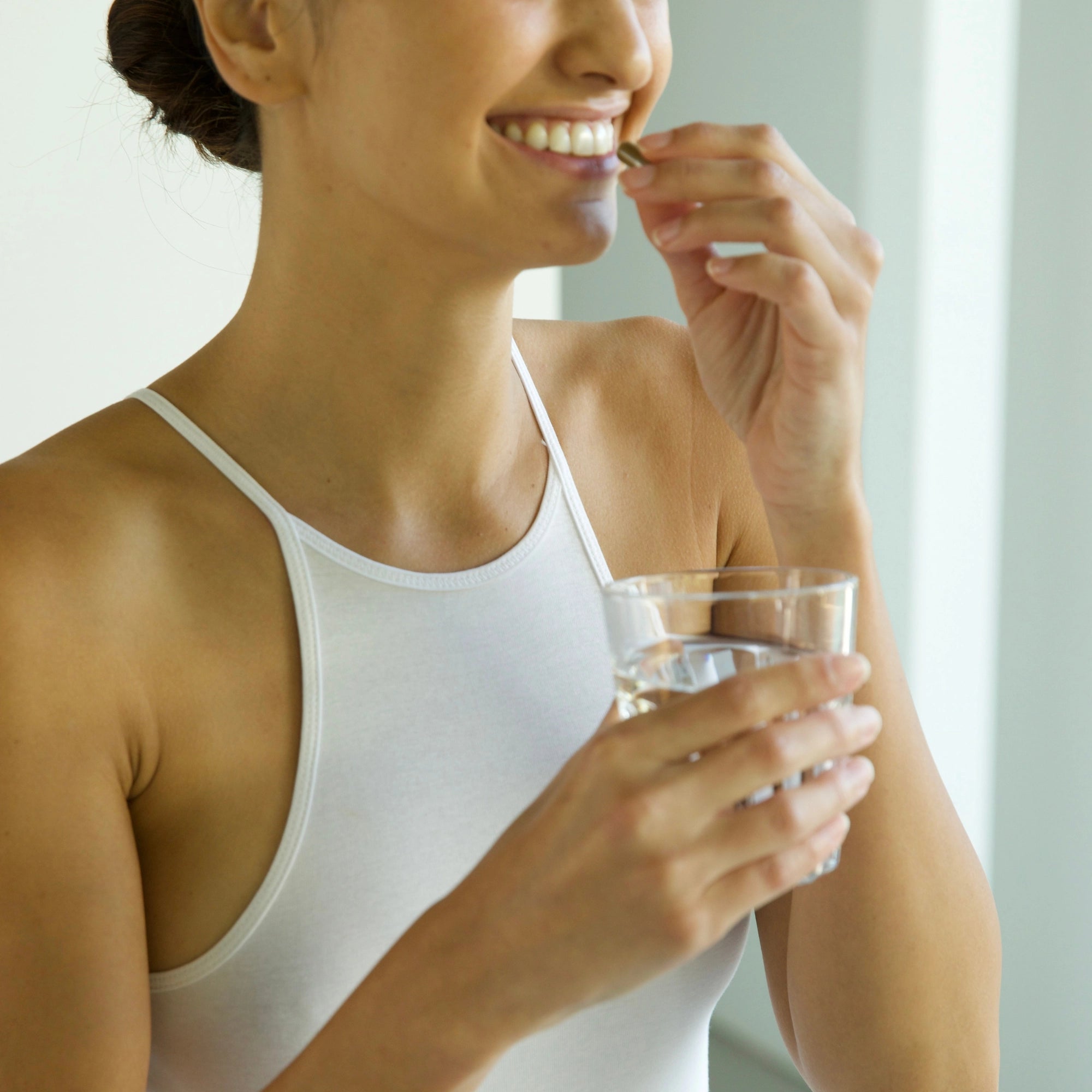 Eine Frau in einem weißen Tank-Top lächelt, während sie ein Glas Wasser in der Hand hält und DALUMA IMMUN DEFENCE COMPLETE KOMPLEX DOUBLE einnimmt, um ihr Immunsystem zu stärken und von seiner antimikrobiellen Wirkung zu profitieren.