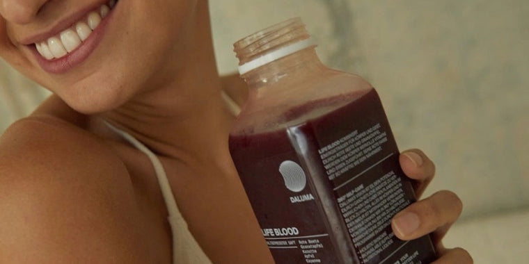 Eine lächelnde Person, die eine Flasche dunkelroten Saft mit einem teilweise sichtbaren Etikett in der Hand hält und ein helles Tanktop trägt.