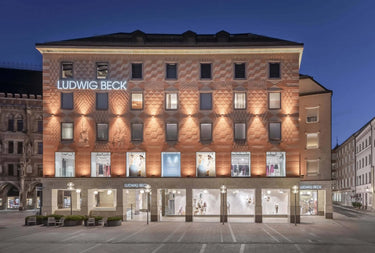 Ein großes, beleuchtetes Gebäude in der Abenddämmerung mit Ludwig Beck-Beschilderung, in dessen Schaufenstern Schaufensterpuppen und Mode ausgestellt sind. Die Architektur zeichnet sich durch gemustertes Mauerwerk und dekorative Statuen über dem Eingang aus.