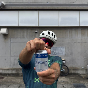 Ein Radfahrer, der einen weißen Helm und eine Sonnenbrille trägt, hält eine Dose DALUMA CLEAN BCAA ENERGY DRINK in die Kamera. Im Hintergrund ist ein Fahrrad an einer Betonwand geparkt.