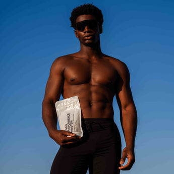 Ein Mann ohne Hemd und mit dunkler Sonnenbrille steht vor einem strahlend blauen Himmel und hält eine weiße Verpackung mit der Aufschrift PERFORMANCE PROTEIN in einer Hand.