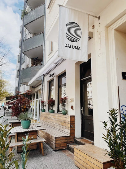 Ein gemütliches Café mit hölzernen Sitzgelegenheiten im Freien und Topfpflanzen liegt an einer Stadtstraße. Über dem Eingang hängt eine weiße Fahne mit dem Logo und dem Namen "DALUMA". In der Nähe sind die Balkone der Wohnungen zu sehen.