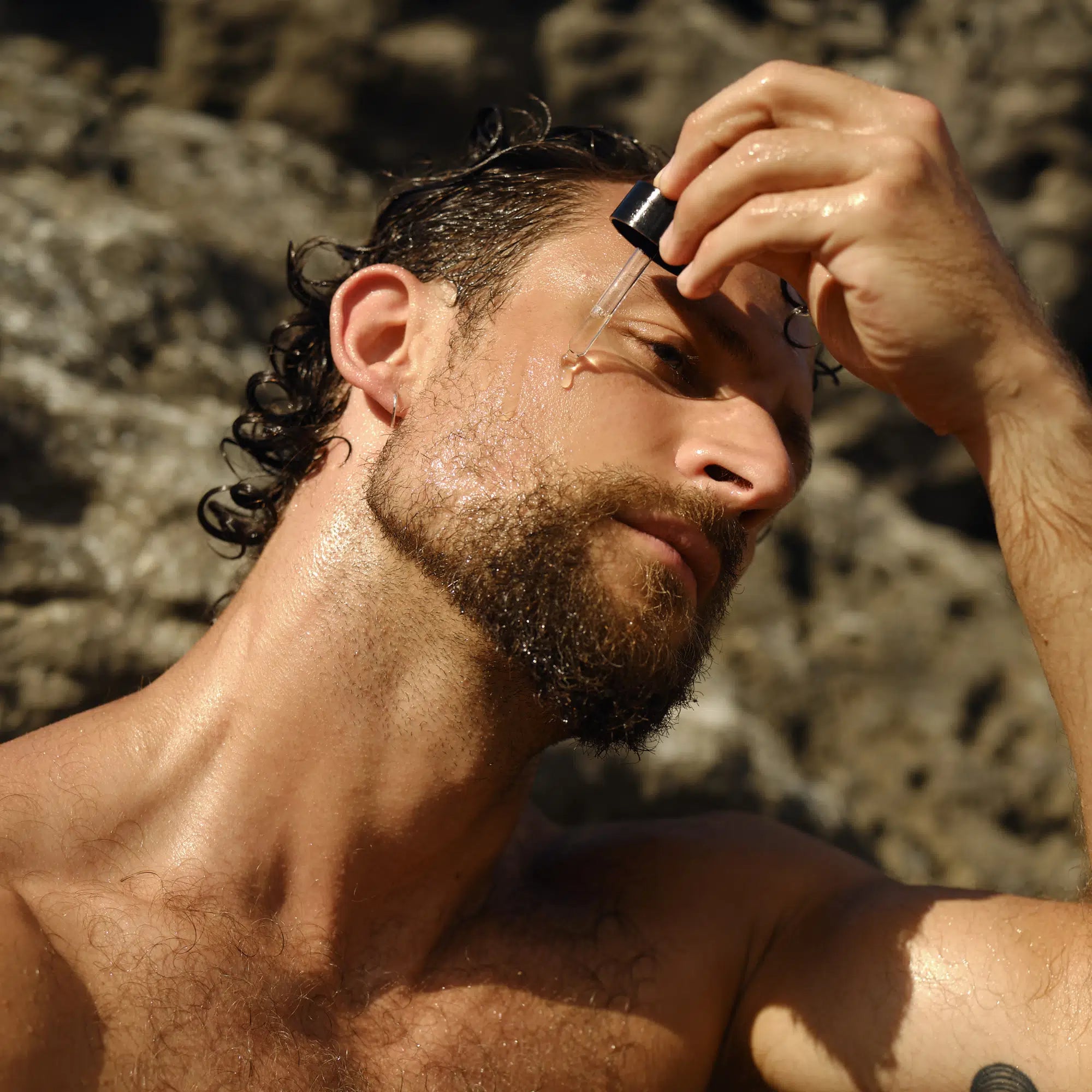 Ein bärtiger Mann trägt DALUMA RADIANT GLAZE GESICHTSÖL mit einer Pipette auf seine Wange auf, während er draußen nahe Felsen steht. Seine Haut erscheint feucht und von der Sonne verwöhnt.