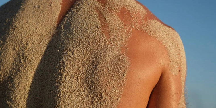 Eine Nahaufnahme des nackten Rückens einer Person, der im Sonnenlicht mit Sand bedeckt ist, vor einem klaren blauen Himmel.