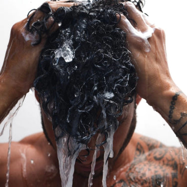 Eine Person mit nassem, lockigem Haar und tätowierten Armen seift sich unter fließendem Wasser mit dem DALUMA STRENGTHENING HAIR WASH FEUCHTIGKEITSSHAMPOO ein, pflegt die Locken und beruhigt die Kopfhaut, während Wasser und Seifenschaum den Kopf hinunterlaufen.