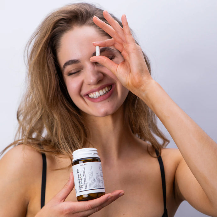Eine lächelnde Frau mit langen Haaren hält eine DALUMA SYM-BIOTIC 23/100 BILLION DARMBAKTERIEN DOUBLE Flasche in der einen Hand und eine Kapsel in der Nähe ihres Auges, vor einem schlichten hellen Hintergrund.