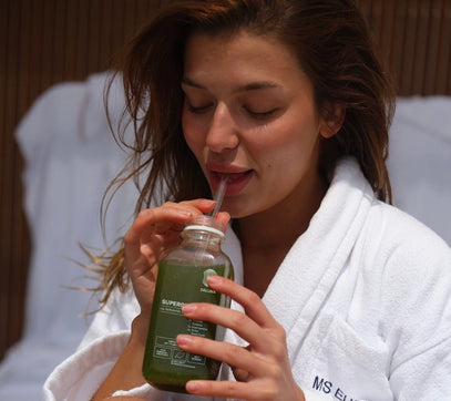 Eine Frau in einem weißen Bademantel trinkt einen grünen Smoothie aus einer Glasflasche mit einem Strohhalm. Sie wirkt entspannt und sitzt auf einem weißen, gepolsterten Stuhl, mit einem leichten Lächeln im Gesicht.