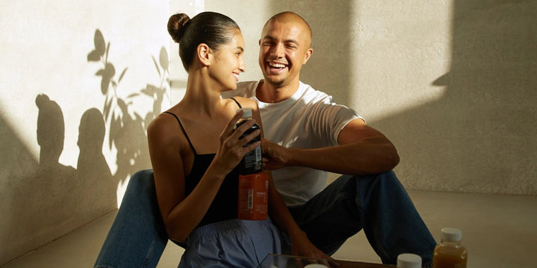 Ein lächelndes Paar sitzt auf dem Boden im Sonnenlicht, hält Getränke in der Hand und ist einander zugewandt. Die Frau trägt ein schwarzes Oberteil und eine hellblaue Hose, der Mann ein weißes T-Shirt. Flaschen stehen in der Nähe auf dem Boden, Schatten werden an die Wand hinter ihnen geworfen.