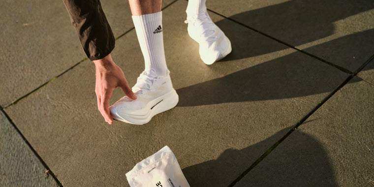 Eine Person, die weiße Adidas-Socken und weiße Laufschuhe trägt, bückt sich auf einer gepflasterten Fläche und greift nach einem weißen Päckchen, das auf dem Boden liegt.