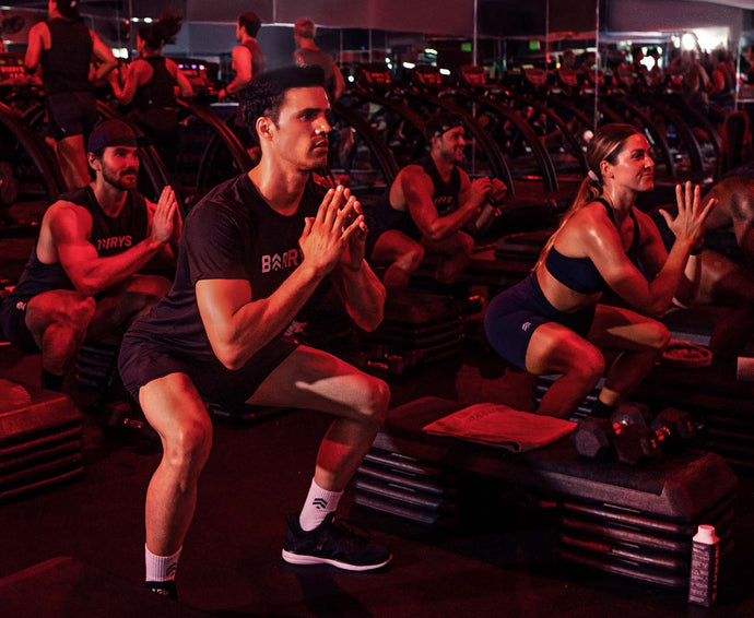 Eine Gruppe von Menschen trainiert in einer schwach beleuchteten Turnhalle und macht Kniebeugen auf Stufenplattformen. Sie tragen Trainingskleidung und wirken konzentriert, wobei die rote Beleuchtung die Szene noch intensiver macht.