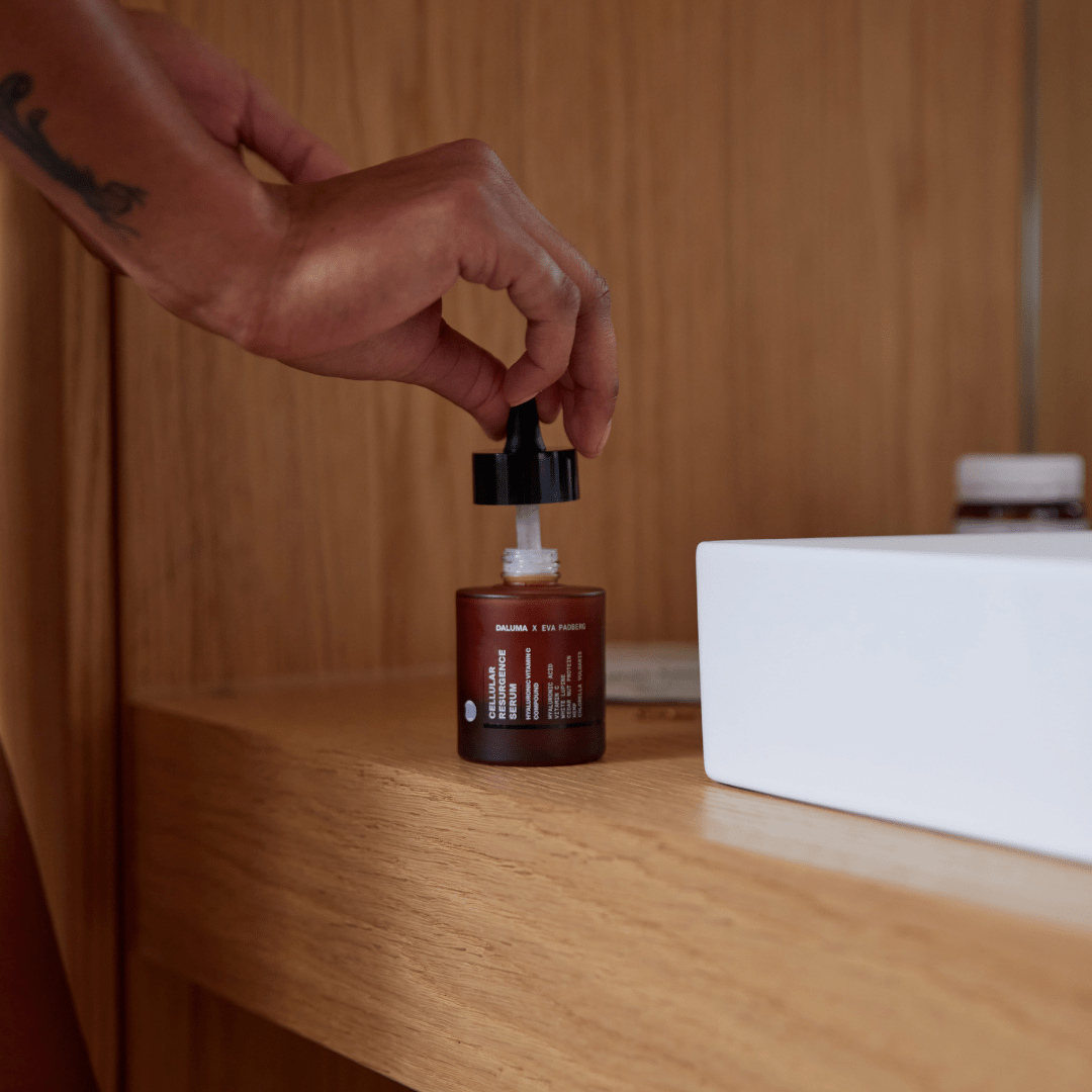 Eine Hand hält den Tropfer von DALUMA's ANTI AGING ESSENTIAL ROUTINE, einer braunen Glasflasche mit schwarzem Verschluss, auf einem Holzregal neben einem weißen rechteckigen Behälter und einer weiteren kleinen Flasche.