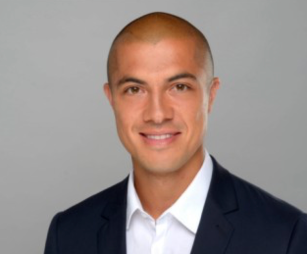 Lächelnder Mann mit rasiertem Kopf, der eine dunkle Anzugsjacke und ein weißes Hemd mit Kragen trägt und vor einem schlichten hellgrauen Hintergrund posiert.