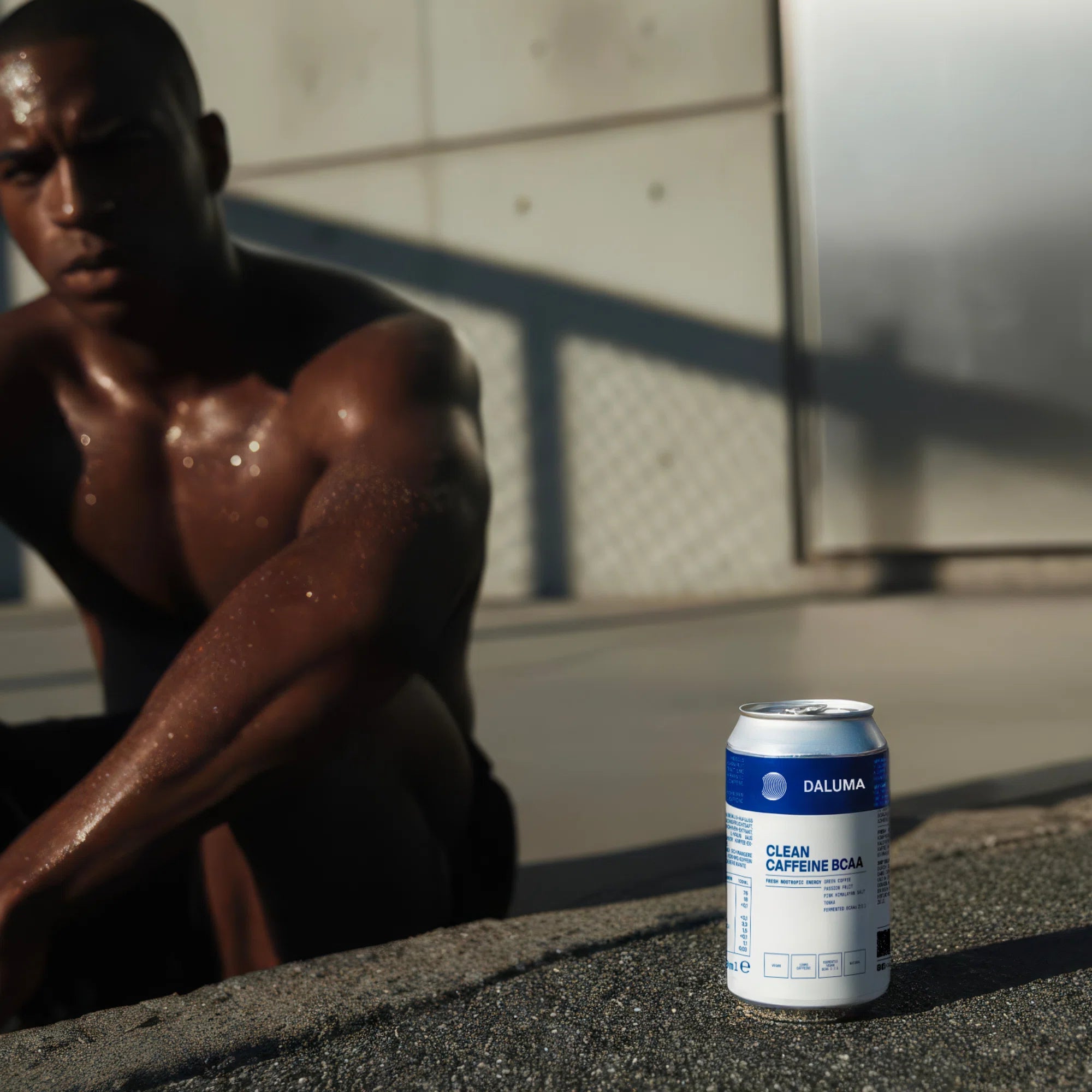 Ein verschwitzter Mann in Trainingskleidung sitzt im Freien im Sonnenlicht, den Arm auf dem Knie. Im Vordergrund ruht eine Dose DALUMA CLEAN BCAA ENERGY DRINK auf dem Beton neben ihm.