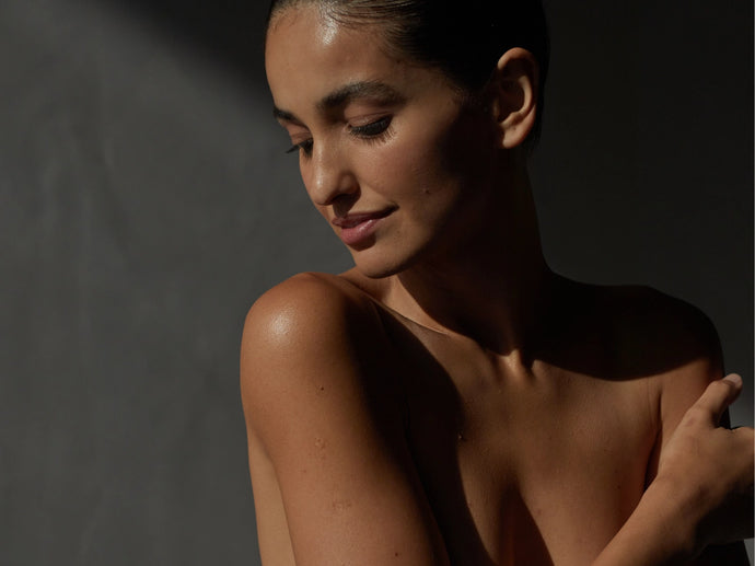 Eine Frau mit hochgestecktem dunklem Haar blickt mit einem sanften Lächeln nach unten, ihre nackte Schulter wird von sanftem Licht vor einem dunklen, minimalistischen Hintergrund beleuchtet.