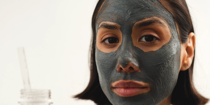 Eine Frau mit glattem dunklem Haar trägt eine graue Gesichtsmaske aus Ton, die ihre Stirn, Nase, Wangen und ihr Kinn bedeckt. Sie blickt direkt in die Kamera und hat einen neutralen Ausdruck. Der Hintergrund ist schlicht und in hellen Farben gehalten.