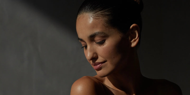 Eine Frau mit glatter Haut und dunklem, zurückgekämmtem Haar blickt mit sanftem Blick nach unten, beleuchtet von weichem, natürlichem Licht vor einem dunklen, neutralen Hintergrund.