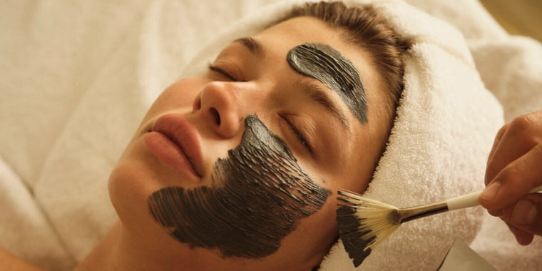 Eine Person, die ein Handtuch um den Kopf gewickelt hat, liegt mit geschlossenen Augen, während jemand mit einem Pinsel eine dunkle Gesichtsmaske auf ihr Gesicht aufträgt.