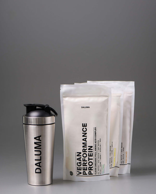 Eine Shakerflasche aus Edelstahl mit der Aufschrift DALUMA steht neben drei weißen Beuteln mit der Aufschrift VEGAN PERFORMANCE PROTEIN, ebenfalls mit der Marke Daluma, vor einem schlichten grauen Hintergrund.