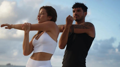 Zwei Personen strecken ihre Arme an einem Strand in den Sonnenuntergang. Die Frau trägt ein weißes Sportoutfit, der Mann ein schwarzes Tanktop und Shorts. Das Meer und eine weitere Person im Wasser sind im Hintergrund zu sehen.