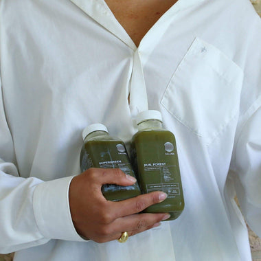 Eine Person in einem weißen Hemd hält zwei Flaschen DALUMA DAILY GREEN JUICE PACKAGE mit weißem Deckel, die reich an Mikronährstoffen sind. Text und Logos sind sichtbar, während ihre Hand die grünen Säfte teilweise umschließt.