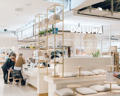 Modernes, minimalistisches Café-Interieur mit weiß gefliesten Oberflächen, Holzakzenten und gepolsterten Sitzgelegenheiten. An der Theke sitzen mehrere Personen. An der Wand hinter der Theke leuchtet das Schild "DALUMA".