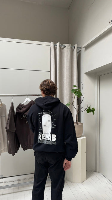 In einem Innenraum steht eine Person, die den JOSEPH ATELIER X DALUMA REHAB HOODIE von DALUMA trägt - einen schwarzen Kapuzenpullover in Übergröße mit weißer Grafik und dem Text REHAB" auf dem Rücken. Ein Kleiderbügel, ein Kleiderständer, eine Topfpflanze und ein Vorhang vervollständigen die Szene.