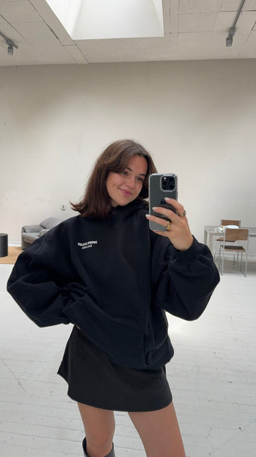 Eine Frau mit schulterlangen braunen Haaren macht ein Spiegel-Selfie in einem hellen, minimalistischen Raum. Sie trägt den schwarzen JOSEPH ATELIER X DALUMA REHAB HOODIE von DALUMA in Übergröße und einen schwarzen Rock. Sie lächelt leicht und stützt eine Hand in die Hüfte.