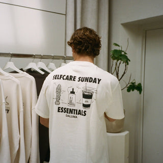 Eine Person mit kurzen, gewellten Haaren ist von hinten zu sehen, die das DALUMA JOSEPH ATELIER X DALUMA SELFCARE SUNDAY TEE trägt, ein übergroßes weißes T-Shirt mit schwarzen Abbildungen und Text. Sie stehen neben einem Kleiderständer und einer kleinen Topfpflanze.