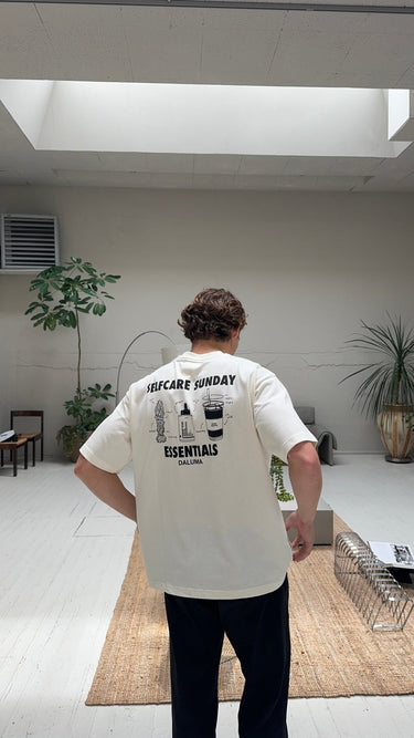 Eine Person mit gelockten Haaren steht in einem Haus, den Blick abgewandt, und trägt das JOSEPH ATELIER X DALUMA SELFCARE SUNDAY TEE von DALUMA. Auf dem übergroßen weißen T-Shirt sind Illustrationen und Text auf dem Rücken abgebildet. Der minimalistische Raum hat helle Wände, Pflanzen und moderne Möbel.