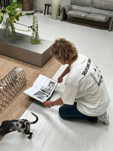 Eine Person, die das DALUMA JOSEPH ATELIER X DALUMA SELFCARE SUNDAY TEE trägt, kniet auf einem weißen Boden und schaut in ein aufgeschlagenes Buch. In dem hellen, minimalistischen Raum mit modernen Möbeln und einer Pflanze läuft ein kleiner Hund vorbei.