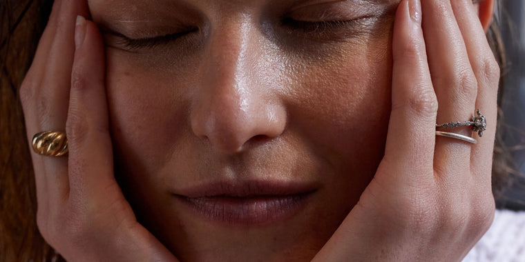 Nahaufnahme einer Person mit geschlossenen Augen, die sich mit beiden Händen sanft das Gesicht hält und zwei Ringe - einen goldenen und einen silbernen - an ihren Fingern trägt. Der Ausdruck wirkt ruhig und nachdenklich.