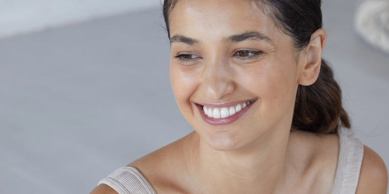 Eine Frau mit zurückgebundenem dunklem Haar lächelt und blickt leicht zur Seite. Sie trägt ein helles, ärmelloses Oberteil und befindet sich in einem schwach beleuchteten Innenraum.