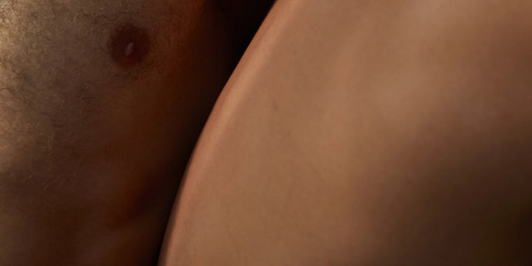 Nahaufnahme zweier Personen, die sich mit ihrer nackten Haut berühren, wobei ein Teil der Brust mit einer Brustwarze und die Seite einer anderen Person zu sehen sind, wodurch eine intime und weiche Komposition entsteht.