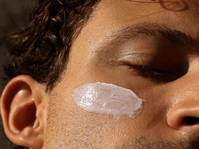 Eine Nahaufnahme des Gesichts einer Person mit geschlossenen Augen, die einen Abstrich weißer Creme auf der Wange hat. Die Beleuchtung hebt die Hautbeschaffenheit und die Gesichtsbehaarung hervor.