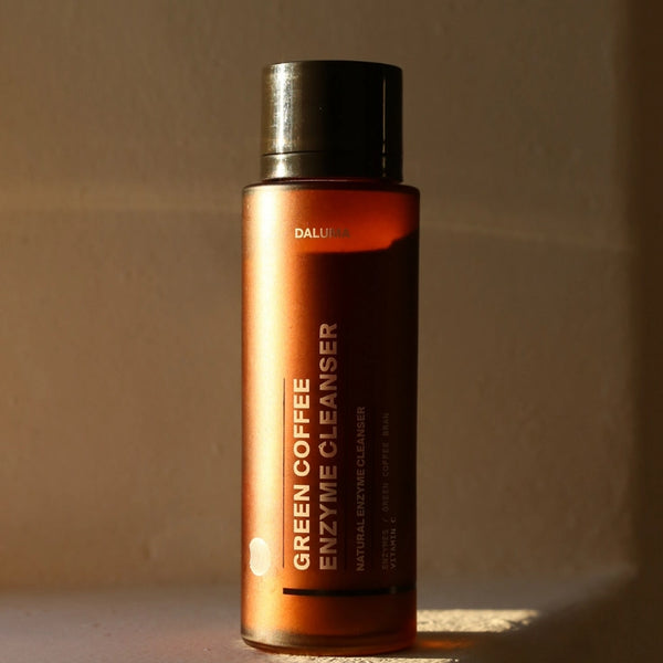 Eine braune Flasche mit der Aufschrift Green Coffee Enzyme Cleanser steht aufrecht auf einer Fläche, die von warmem Sonnenlicht beleuchtet wird. Der Verschluss ist schwarz, und die Flasche ist mit minimalistischem Text und Design versehen.