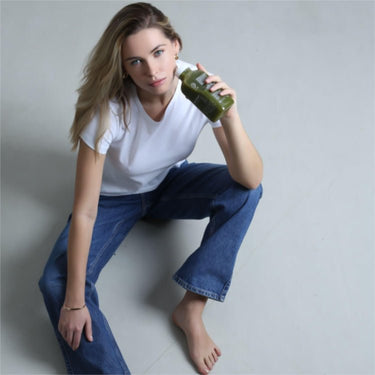 Eine Frau mit langen blonden Haaren, barfuß, in blauen Jeans und weißem T-Shirt, sitzt auf dem Boden und schaut mit einem Glas DALUMA GRÜNER SÄFTIG PLUS SAFTKUR 3 TAGE in die Kamera.